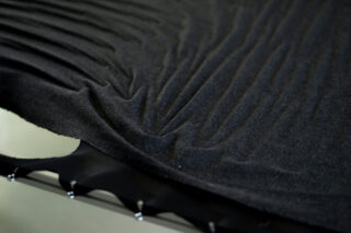 Nahaufnahme eines strukturierten schwarzen Stoffes mit sichtbaren Falten und Knicken, der über eine Oberfläche mit Metallzähnen oder -zinken am Rand gespannt ist.
