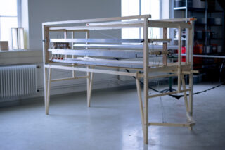 Ein großer Holzwebstuhl mit Metallstäben und -fäden steht in einem gut beleuchteten Raum mit weißen Wänden und Industrieregalen.