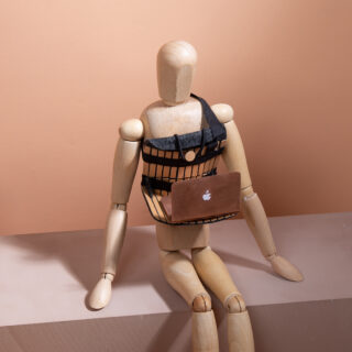 Eine hölzerne Schaufensterpuppe sitzt mit einem kleinen Laptop auf einem Sims und trägt vor einem schlichten beigen Hintergrund ein schwarzes, geschirrähnliches Accessoire.