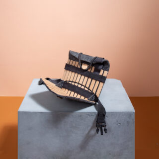 Eine rechteckige Tasche aus Lattenholz und schwarzem Stoff ruht auf einem grauen Würfel vor einem zweifarbigen beige-braunen Hintergrund.