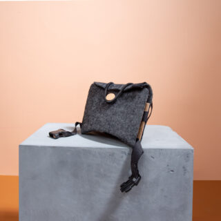 Eine rechteckige graue Filztasche mit Knopfverschluss und verstellbaren schwarzen Riemen steht auf einem grauen Sockel vor einem pfirsichfarbenen Hintergrund.