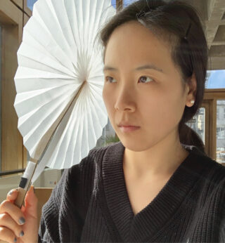 Eine Person in einem schwarzen Pullover hält einen weißen Papierfächer, im Hintergrund scheint Sonnenlicht durch ein Fenster.