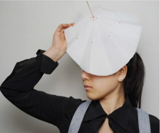 Eine Person mit schwarzem Hemd und grauen Hosenträgern hält einen kleinen, weißen, gefalteten Papierschirm über den Kopf und bedeckt so teilweise ihr Gesicht.