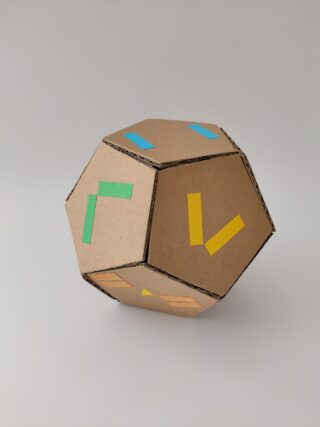 Vor einem schlichten Hintergrund wird ein Dodekaeder aus Pappe mit farbigen L-förmigen Aufklebern auf jeder Seite angezeigt.