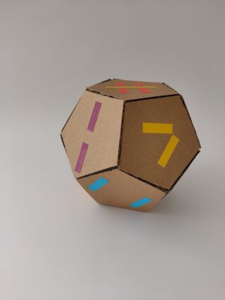 Ein Dodekaeder aus Pappe mit farbigen Klebestreifen und Zahlen auf den Flächen liegt auf einer einfachen Fläche vor einem neutralen Hintergrund.