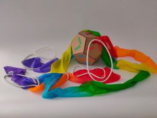 Ein Papppolyeder mit farbigem Klebeband liegt neben zwei lila Schlafmasken und einem langen, regenbogenfarbenen Schal auf einem schlichten Hintergrund.