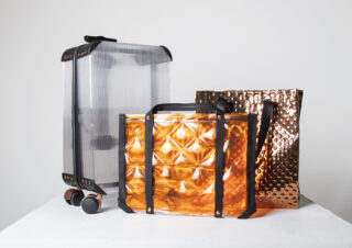 Drei moderne Taschen aus metallischen Materialien: ein durchscheinender Koffer mit schwarzen Riemen, eine orangefarbene Tragetasche mit geometrischem Muster und eine strukturierte Tasche in Kupfertönen, alle auf einer weißen Oberfläche platziert.