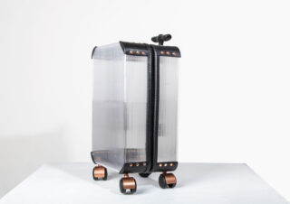 Ein transparenter Hartschalenkoffer mit schwarzem Rand und Kupferakzenten steht aufrecht auf vier Rädern vor einem schlichten Hintergrund.