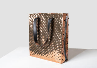 Auf einer schlichten weißen Fläche liegt eine metallische, kupferfarbene Tragetasche mit strukturierter, genoppter Oberfläche und zwei schwarzen Henkeln.