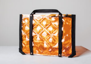 Eine rechteckige, orangefarbene, durchscheinende Tasche mit Steppmuster und schwarzen Griffen liegt auf einer weißen Fläche vor einem hellgrauen Hintergrund.