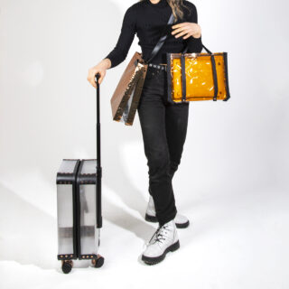 Eine Person in schwarzem Outfit hält vor einem schlichten weißen Hintergrund gelbe und braune transparente Taschen und zieht einen durchsichtigen Hartschalen-Rollkoffer.