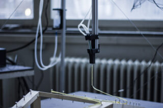 Ein Roboterarm mit einem dünnen Rohraufsatz arbeitet in einer Laborumgebung über einem Holzrahmen mit Nägeln.