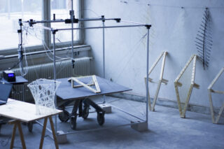 In einer industriellen Werkstatt wird auf einem Tisch mithilfe eines großen 3D-Druckers eine geometrische Struktur konstruiert. In der Nähe befinden sich Modelle und Holzrahmen.