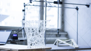 Eine durchscheinende, geometrische 3D-gedruckte Struktur steht auf einem Holztisch in einem Arbeitsbereich mit technischen Geräten und einem Laptop im Hintergrund.