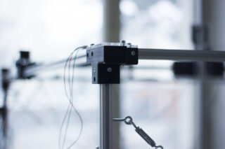 Nahaufnahme einer Metallrahmenstruktur mit daran befestigten Drähten, mit schwarzen Eckverbindungen und Bolzen, vor einem unscharfen Innenhintergrund.