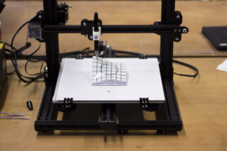 Ein 3D-Drucker druckt ein weißes Objekt mit einem schwarzen Gittermuster auf einem Flachbett, das auf einem Holztisch steht.
