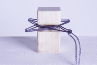 Ein aufrecht stehender Holzblock ist mit einer violetten Schnur umwickelt und festgebunden, sodass auf der Vorderseite eine sternförmige Form entsteht.