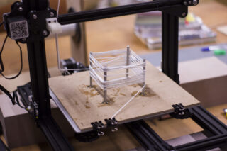 Ein 3D-Drucker im Betrieb, der mit weißem Filament auf einer flachen Bauplattform eine kleine quadratische Rahmenstruktur erstellt.