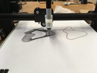 Ein Plotter im Stil eines 3D-Druckers zeichnet abstrakte schwarze Linien auf ein weißes Blatt Papier und erstellt so auf seiner flachen Arbeitsfläche ein geometrisches Design.