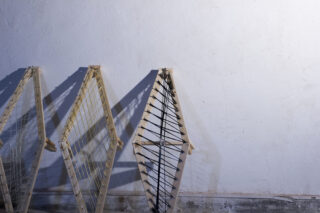 Drei Holzrahmen mit Drahtgeflecht und Schnurmustern lehnen an einer weiß strukturierten Wand auf einem Betonboden.