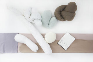 Verschiedene weiche, stoffbezogene Kissen und Nackenrollen in neutralen Farbtönen sind auf einer flachen Fläche neben einem Skizzenbuch mit einfachen handgezeichneten Formen angeordnet.