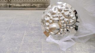 Ein rundes, silbernes, strukturiertes Metallobjekt mit erhabenen, blasenartigen Formen liegt teilweise in durchscheinenden Kunststoff eingewickelt auf einer gefliesten Oberfläche.