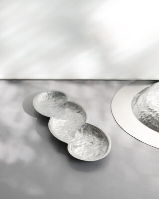 Drei runde, strukturierte Silberschalen sind in einer Reihe miteinander verbunden und stehen auf einer glatten, hellgrauen Oberfläche mit gesprenkeltem Licht und Schatten.