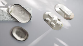 Metallische Tabletts mit erhabenen, strukturierten Oberflächen sind auf einer hellen Fläche neben einem Granitblock angeordnet; auf einem Tablett liegt eine kleine Blume.