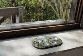 Eine Keramikfarbpalette mit getrockneter Farbe steht auf einem Fensterbrett neben einem offenen Fenster, draußen sind grüne Pflanzen zu sehen.