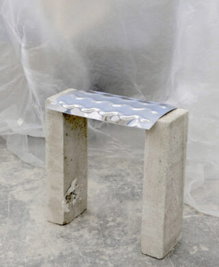 Zwei Betonblöcke stehen aufrecht, auf deren Oberseite ein gewelltes Metallblech angebracht ist, und bilden so eine minimalistische Bank oder einen Hocker. Im Hintergrund hängt eine durchscheinende Kunststoffplatte.