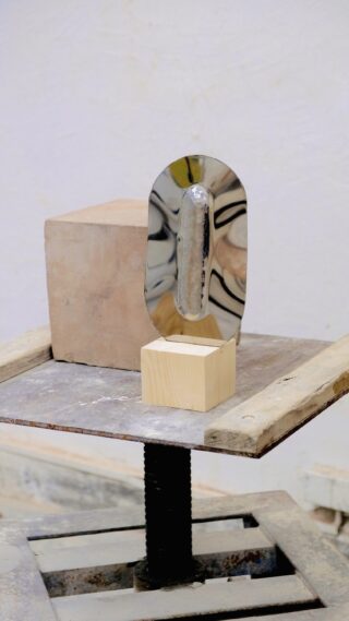 Eine Metallkelle steht aufrecht an einem beigen Ziegelstein und ein kleiner Holzblock steht auf einer Metallwerkbank mit einem Schraubstock.