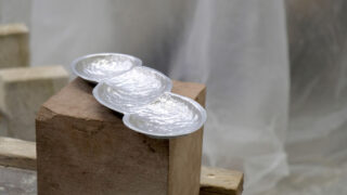 Drei verbundene Silberschalen mit strukturierten Oberflächen ruhen auf einem groben rechteckigen Holzblock vor einem unscharfen, neutralen Hintergrund.