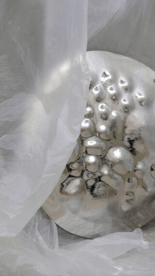 Ein silbernes, strukturiertes Metallobjekt, das teilweise mit einem durchsichtigen, zerknitterten weißen Stoff bedeckt ist.