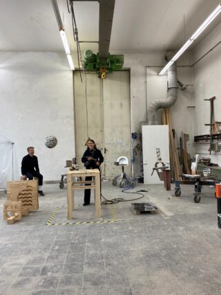 Zwei Personen in einer Werkstatt mit Holzbearbeitungswerkzeugen, Geräten und Holzskulpturen; eine steht neben einem Tisch, während die andere auf einem Holzstück sitzt.