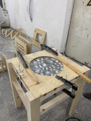 Ein Holzrahmen hält ein Metallblech mit genoppter Oberfläche, das mit mehreren Klammern auf einem Werkstatttisch befestigt ist; im Hintergrund sind verschiedene Holzobjekte zu sehen.