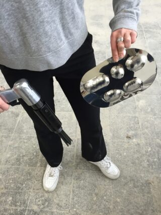 Eine Person in einem grauen Sweatshirt und einer schwarzen Hose hält in der einen Hand einen metallischen Gegenstand mit Rillen und in der anderen ein Gerät mit Griff und Düse und steht auf einem gefliesten Boden.