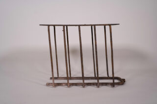 Rostiges Metallgitter mit sechs vertikalen Stäben und einer flachen Oberseite, aufrecht vor einem schlichten weißen Hintergrund positioniert.