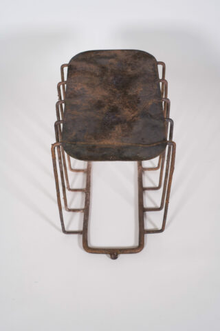 Ein Metallhocker mit rechteckiger, verrosteter Sitzfläche und geometrischen, eckigen Beinen auf weißem Hintergrund.