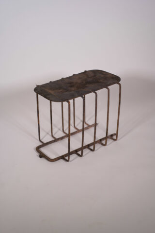 Ein rechteckiges Metallobjekt mit einer flachen, verrosteten Oberseite und sechs gebogenen Drahtbeinen, fotografiert vor einem schlichten, hellen Hintergrund.