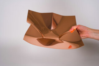 Eine Hand hält ein dünnes, flexibles, glänzendes kupferfarbenes Metallblech, das vor einem schlichten Hintergrund teilweise gefaltet und gebogen ist.