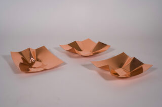 Drei quadratische, metallische, kupferfarbene Schalen mit unregelmäßigen, zerknitterten Formen stehen auf einer schlichten, hellen Fläche vor einem neutralen Hintergrund.