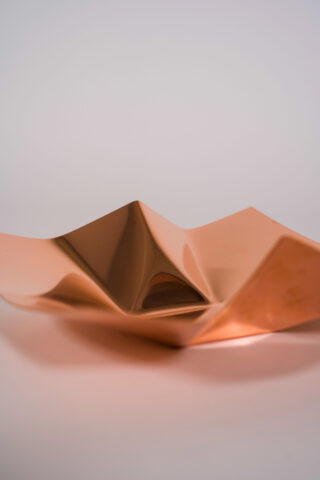 Eine geometrische, eckige Metallschale mit Kupferoberfläche wird auf einer schlichten hellen Oberfläche vor einem neutralen Hintergrund platziert.