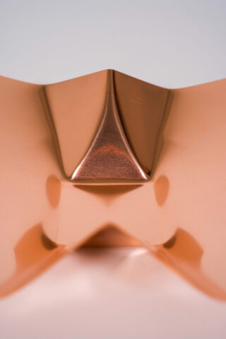 Eine Nahaufnahme eines glatten, gefalteten kupferfarbenen Metallblechs vor einem neutralen Hintergrund.