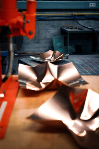 Nahaufnahme von glänzenden, gefalteten Metallblechen auf einer Werkbank in einer Industriewerkstatt, im Hintergrund sind Maschinen und Werkzeuge sichtbar.