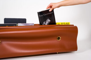 Eine Hand legt ein Buch mit dem Titel „Der Handwerker“ in ein braunes Bücherregal mit gewellten Kanten, in dem Zahnräder und andere Bücher sichtbar sind.