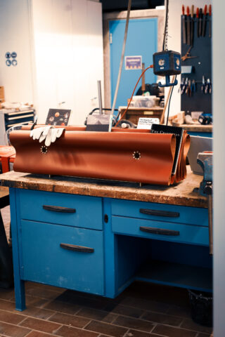 Eine große Kupferplatte mit Ausschnitten liegt auf einer blauen Werkbank in einer Werkstatt, umgeben von Werkzeugen, Handschuhen und Schränken.