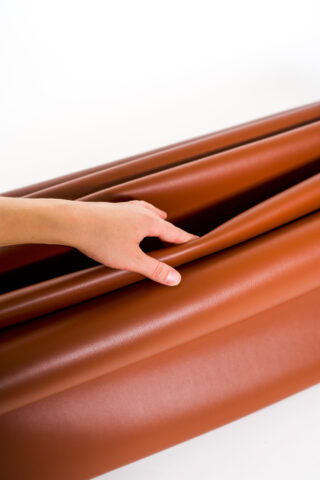 Eine Hand hebt vorsichtig eine Falte aus braunem, lederähnlichem Material an und zeigt so vor einem weißen Hintergrund seine Flexibilität und glatte Textur.