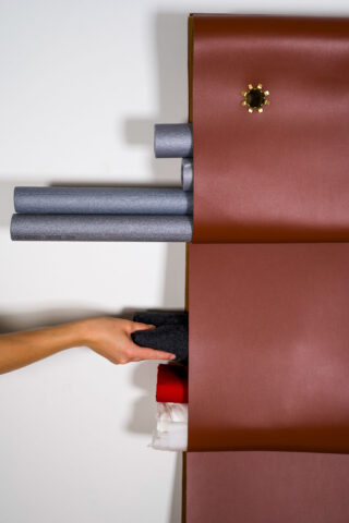 Eine Hand greift nach gestapelten Stoffrollen zwischen zwei großen braunen Stoffbahnen, an deren oberer Bahn eine blumenförmige Nadel befestigt ist.