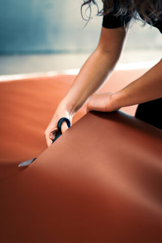 Eine Person schneidet mit einer Schere ein großes Stück braunen Stoff auf einer flachen Oberfläche aus.