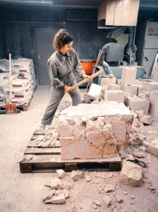 Eine Frau im Overall zertrümmert mit einem Vorschlaghammer einen Steinblock auf einer Palette in einer Industriewerkstatt.